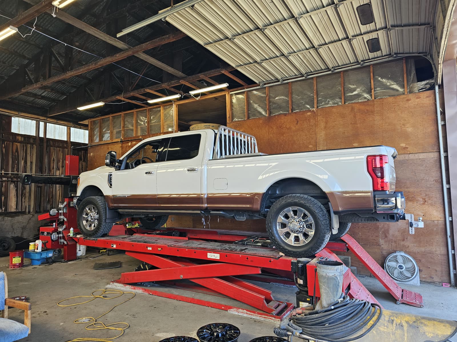 A white and brown Ford F-250 Super Duty up on the red alignment rack inside the timber-framed shop.