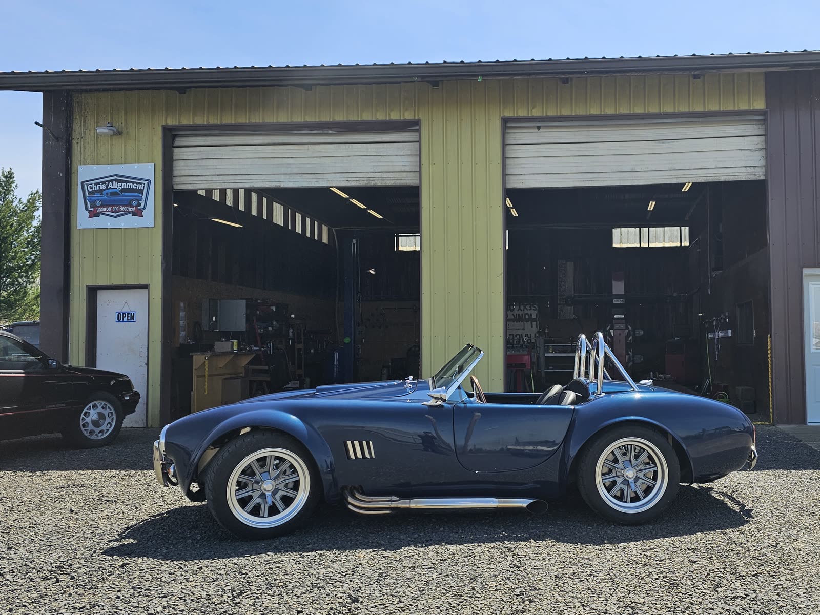 A blue AC Cobra parked on the gravel outside the shop, the Chris' Alignment sign on the wall and both bay doors open.